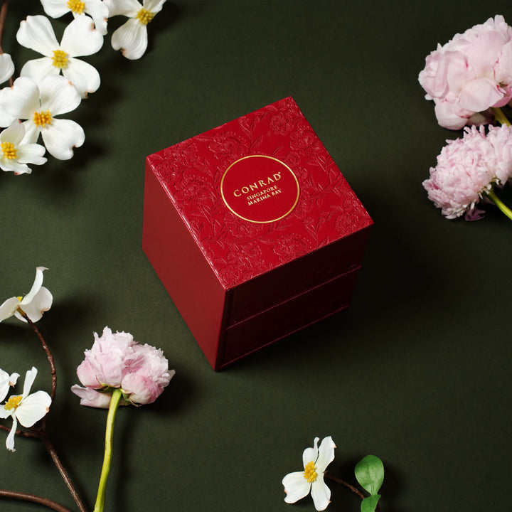 Red jewelry box with 'Conrad' branding surrounded by flowers on a dark background