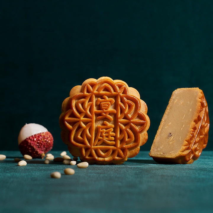 Two mooncakes on a dark teal background, one whole with ornate embossed Chinese characters and the other sliced to show the filling. A lychee and scattered soybeans are placed beside them as styled props.