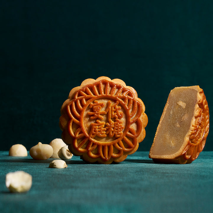 Two mooncakes on a dark teal surface with a few nut pieces scattered in the foreground. One mooncake is whole, showing embossed Chinese characters, and the other is sliced open to show the filling.