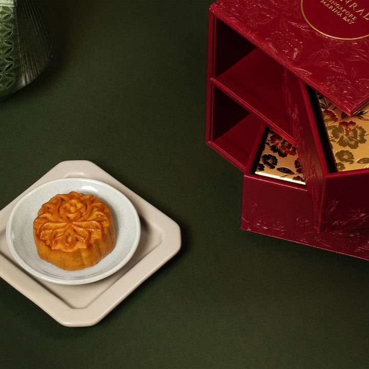 Mooncake on a white plate with an open red gift box containing more mooncakes on a dark green background