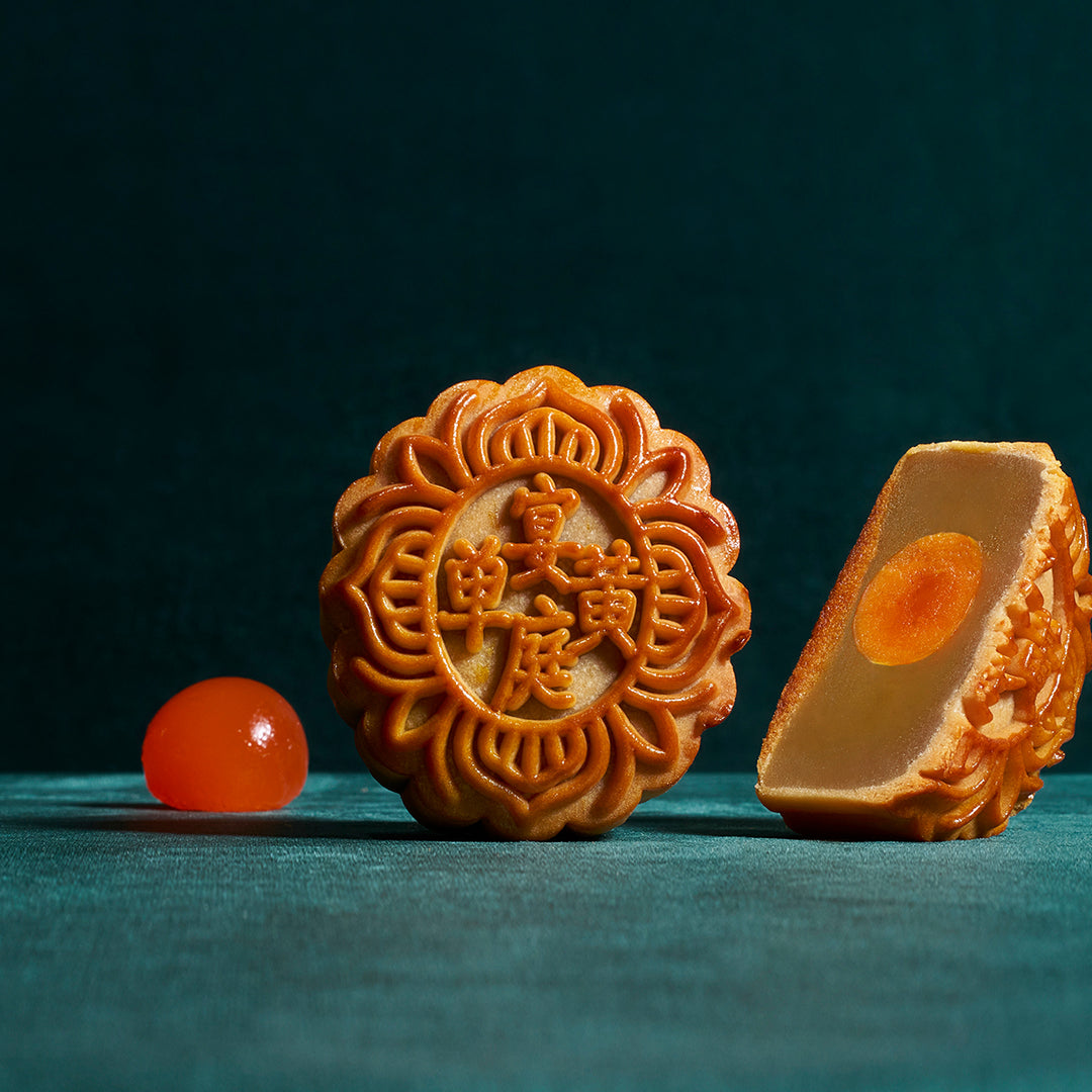 Two mooncakes on a dark teal background, one whole with embossed Chinese characters and one sliced open showing a rich orange filling, with a small orange sphere in the foreground.