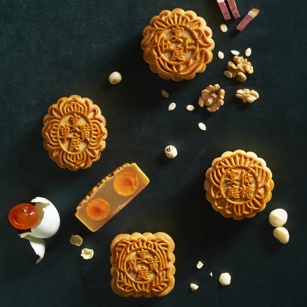 Four Chinese mooncakes with floral embossed patterns are arranged on a dark textured background, alongside a cracked eggshell with red jam, peanuts, and hazelnuts.