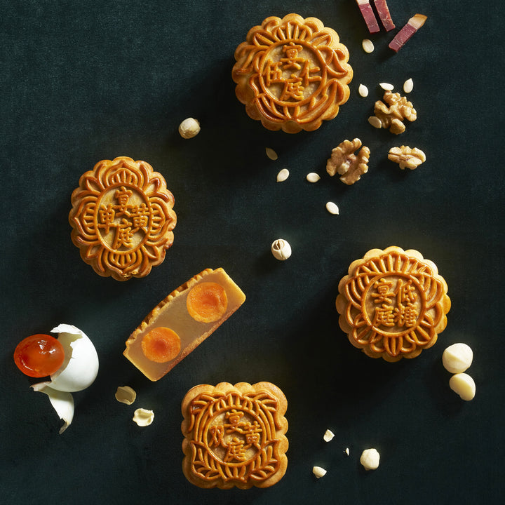 Four Chinese mooncakes with floral embossed patterns are arranged on a dark textured background, alongside a cracked eggshell with red jam, peanuts, and hazelnuts.