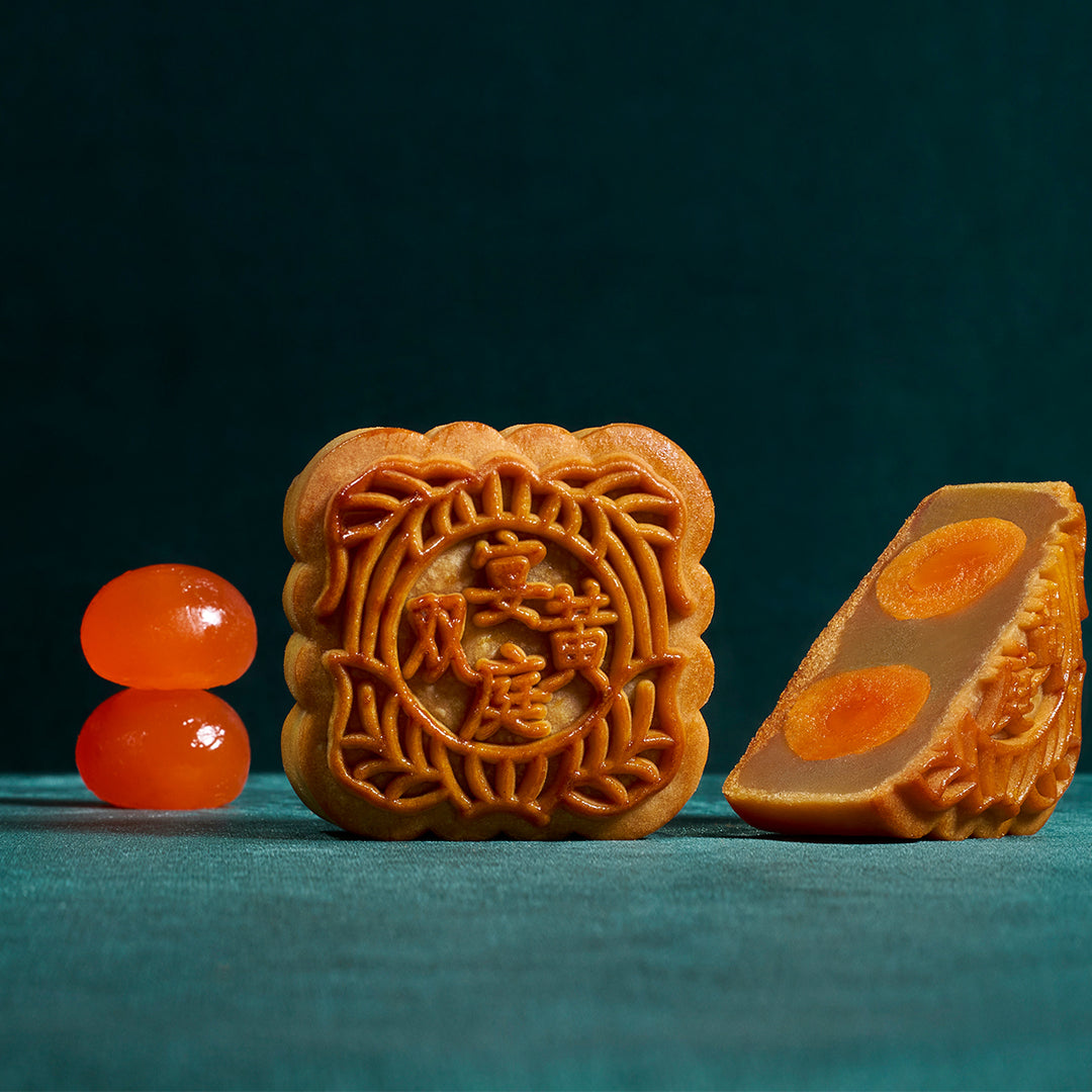 Two traditional Chinese mooncakes on a dark teal background, one whole square mooncake showing embossed Chinese characters and floral patterns, and one sliced mooncake slice showing orange filling. Two glossy orange candied fruit pieces are placed to the left.
