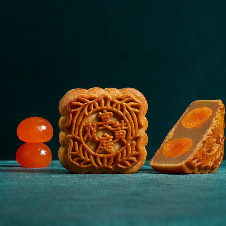 Two traditional Chinese mooncakes on a dark teal background, one whole square mooncake showing embossed Chinese characters and floral patterns, and one sliced mooncake slice showing orange filling. Two glossy orange candied fruit pieces are placed to the left.
